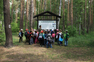 Fotografia przedstawia grupę dzieci, która wraz z przewodnikiem przygląda się tablicy informacyjnej położonej w lesie. Zdjęcie pochodzi z archiwum beneficjenta.