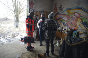 Fotografia przedstawia sytuację w opuszczonym miejscu, gdzie przebywają bezdomni. Widoczne są trzy osoby – dwójka bezdomnych oraz ratowniczka medyczna w pomarańczowym, odblaskowym uniformie, pytająca, czy potrzebna jest pomoc. Autorem zdjęcia jest Dominik Wójcik.