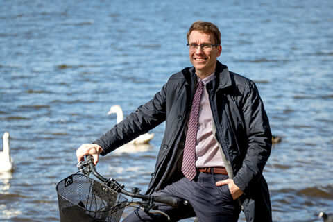 Fotografia przedstawia Krzysztofa W. Jaworowskiego, burmistrza Trzcianki. Młody mężczyzna w okularach, pod krawatem, pozuje na rowerze, na tle jeziora. Zdjęcie pochodzi z archiwum prywatnego.